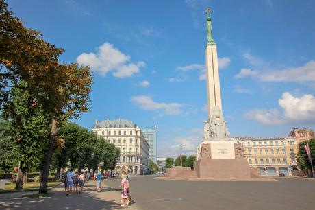 The Freedom Monument in Riga, Latvia, stands tall against a bright blue sky, with trees and historic buildings in the background and people walking nearby.