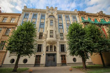 The elaborate facade of an Art Nouveau building, with sculpted details, large windows, and lush green trees flanking the entrance.