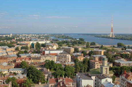 A panoramic view of Riga, Latvia, with a mix of historic and modern buildings, green spaces, and the iconic TV Tower standing tall near the Daugava River.