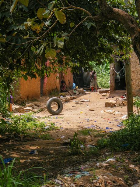 A shaded, dirt path in a rural village, with a large tree overhead and a stray tire in the middle of the path. A child stands near a doorway in the background, and a few colorful houses are partially visible.