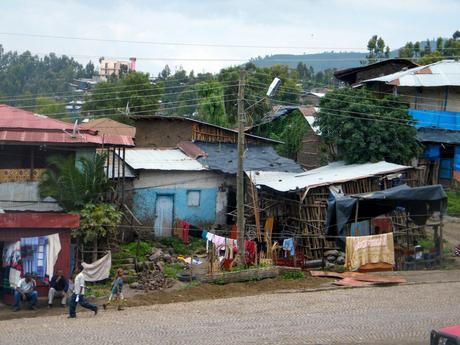 A cluster of small, rustic houses made of wood and corrugated metal, with laundry hanging outside and people going about their daily activities on a cobblestone road.