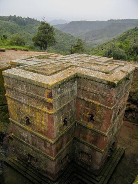 The top view of a large, cross-shaped rock-hewn church, partially submerged in the ground, surrounded by greenery and hills in the background.