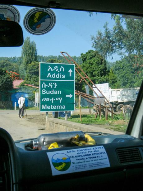 A road sign showing directions to Addis, Sudan, and Metema, viewed from inside a vehicle. The sign is written in both Amharic and English, and the surrounding area includes trees and a person standing nearby.