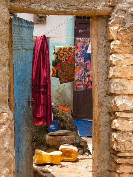 A colorful assortment of clothes hanging on a clothesline in a small, open courtyard with rough stone walls, a blue door, and various household items scattered around.