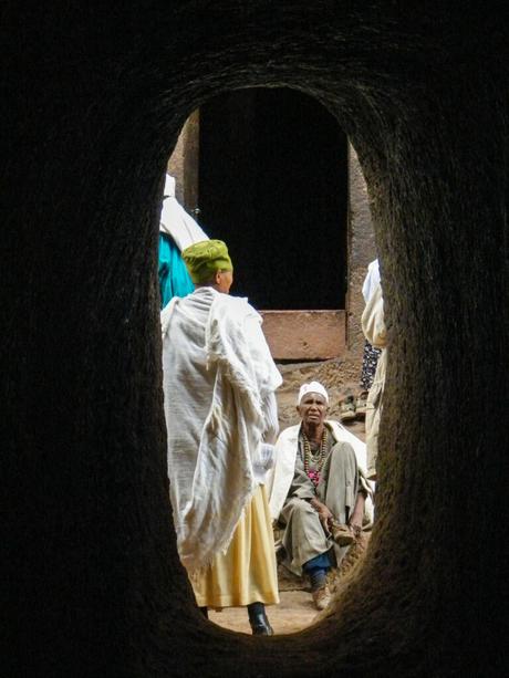 Two Ethiopian priests standing in a rock-hewn church, dressed in white traditional robes, one sitting on the ground and the other standing, viewed through a narrow stone archway.