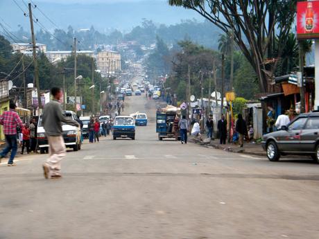 A busy street in an urban area, filled with cars, pedestrians, and small shops lining the sides. People are walking in various directions, and a few vehicles are parked or moving along the road.