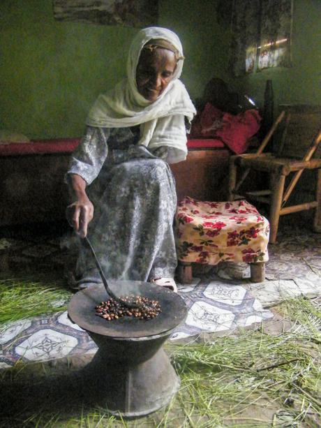 An elderly woman wearing a white headscarf and patterned dress, roasting coffee beans in a traditional Ethiopian coffee ceremony inside a modest room with a few simple furnishings.