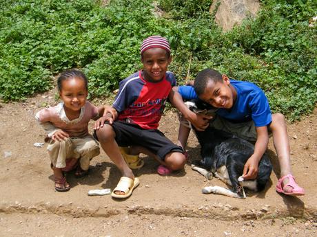 Three smiling children sitting on the ground next to a small black goat, with greenery in the background. The boy in the middle wears a red and black shirt, and the other two children are holding the goat affectionately.