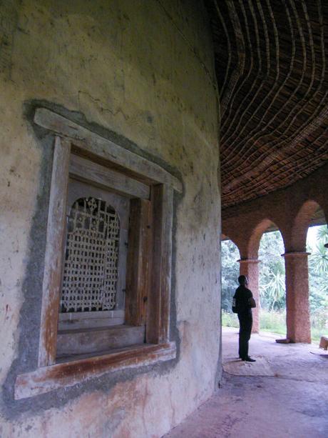 The exterior of a circular building with a thatched roof, showing a wooden window with a mesh covering and a person standing under the arches, silhouetted against the bright outdoors.