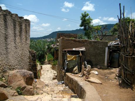 A narrow dirt path between simple stone houses, leading toward green bushes in the background under a clear blue sky.