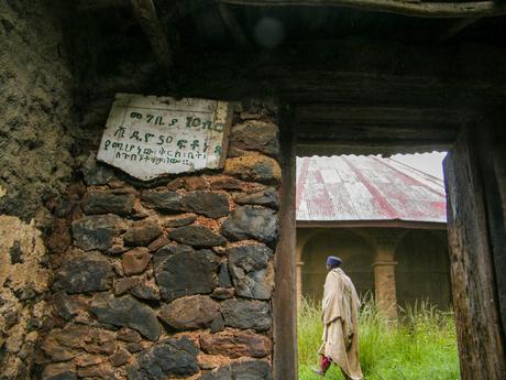 A close-up of a stone wall with a weathered sign in Amharic script, and a person in traditional clothing walking past an open doorway toward a building with a corrugated metal roof.