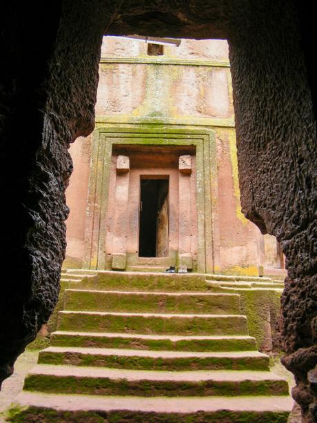 The interior view of a rock-hewn church with a rough, arch-shaped entrance leading to stone steps and an open doorway, with natural light filtering through.