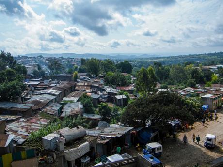 A wide view of a densely packed neighborhood with numerous corrugated metal roofs, surrounded by trees and greenery. The sky is cloudy, with hills visible in the distance.