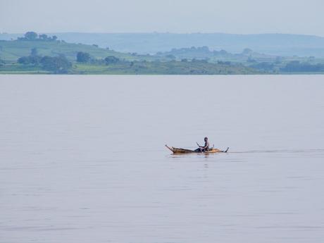 A lone person paddling a small wooden boat on a vast lake, with a distant view of green hills and trees along the shoreline under a hazy sky.