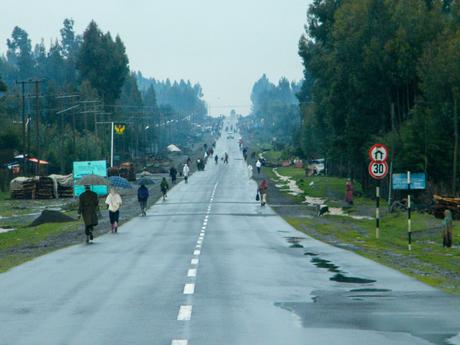 A long, wet road stretching into the distance, with several people walking along both sides under a cloudy sky. Some are carrying umbrellas, and the road is flanked by green trees and small roadside structures.