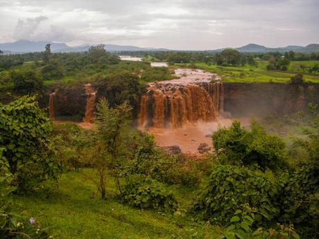 A wide view of a large, muddy waterfall cascading over a rocky cliff, surrounded by lush green vegetation and hills in the distance under a cloudy sky.