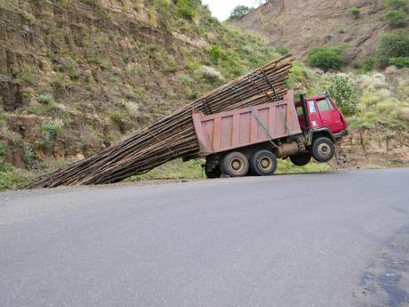 A red truck precariously tilted on a road with its front end lifted in the air due to an overhanging load of long wooden logs, set against a rocky hillside.