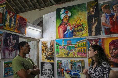 Two people standing in front of a wall filled with colorful paintings, engaged in conversation inside an art gallery.