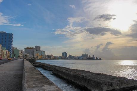 A coastal scene of the Malecón seawall in Havana, Cuba, with the city skyline and the ocean under a partly cloudy sky at sunset.