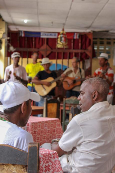 A group of musicians performing in a small venue with guitars and percussion instruments, while two men sit at a table in the foreground, partially in conversation.