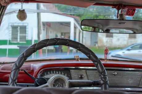 The interior of an old car, showing a cracked steering wheel, a small figure on the dashboard, and a blurred view of a street outside.