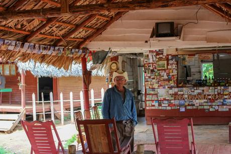 An elderly man wearing a cowboy hat and blue shirt stands inside a rustic wooden hut decorated with postcards and pictures, under a thatched roof.