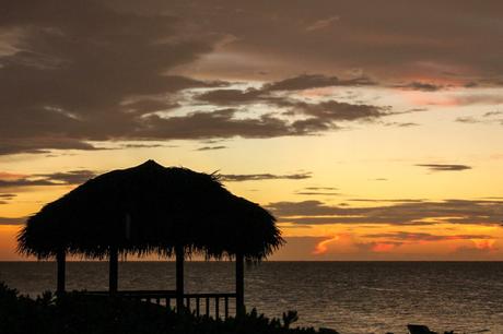 A silhouette of a thatched-roof gazebo against a colorful sunset sky over the ocean.