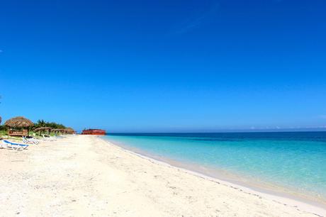 A beautiful sandy beach with turquoise waters, clear blue skies, and several thatched umbrellas along the shore.