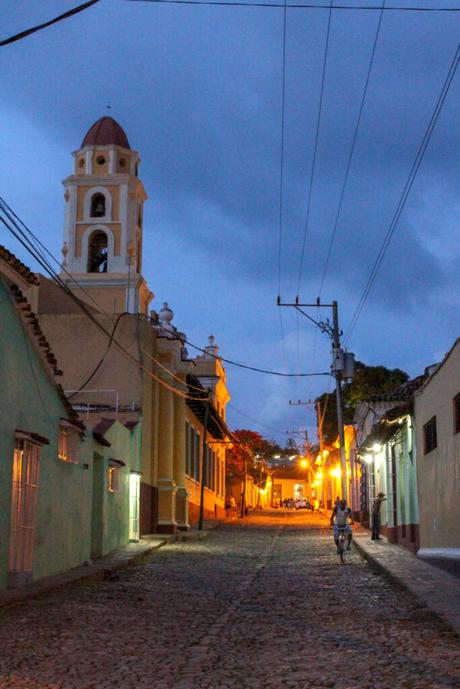 A cobblestone street at dusk with street lamps illuminating colonial-style buildings, including a bell tower in the background.