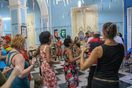 A group of people dancing energetically in a festive room decorated with colorful streamers, with portraits of Che Guevara hanging on the wall in the background.