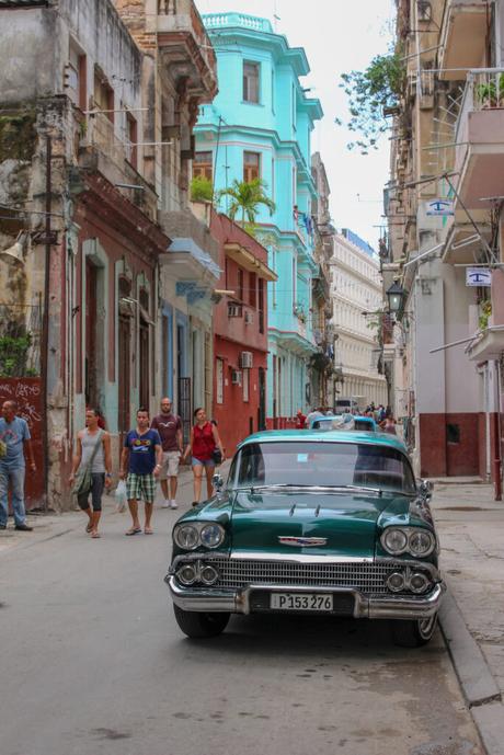 A vibrant street scene featuring a classic green car parked along a narrow street with colorful, weathered buildings, and people walking by.