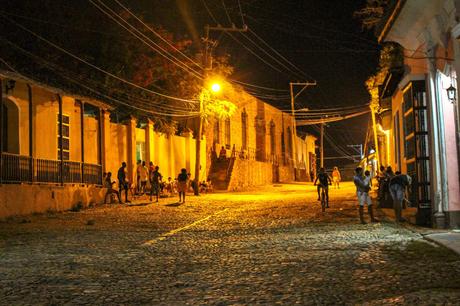 A cobblestone street at night illuminated by yellow streetlights, with people walking and sitting along the sidewalks, and old colonial buildings in the background.