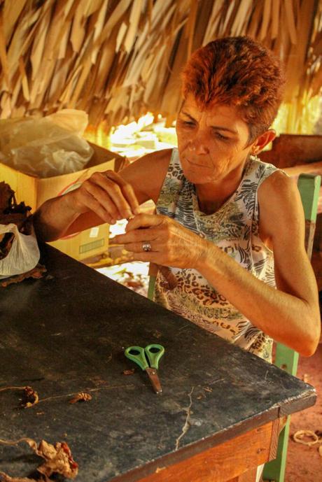 A woman with short red hair sitting at a wooden table, skillfully rolling a cigar inside a rustic tobacco workshop.