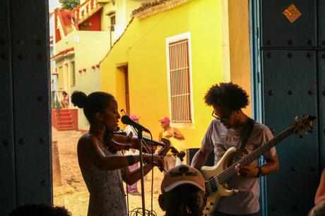 A woman playing a violin and a man playing a bass guitar in an open doorway, with brightly painted yellow and red houses visible outside.