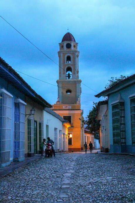 A twilight scene of a cobblestone street with colonial-style buildings and a bell tower illuminated against a dusky blue sky.