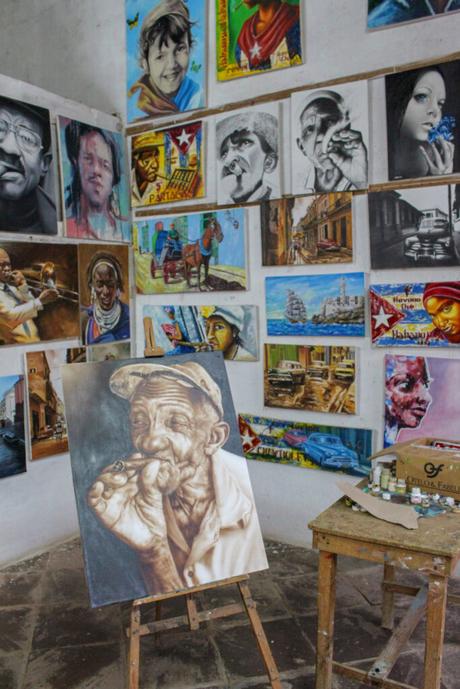 An art studio interior with numerous portraits and paintings on the walls, featuring vibrant depictions of Cuban people and street scenes.