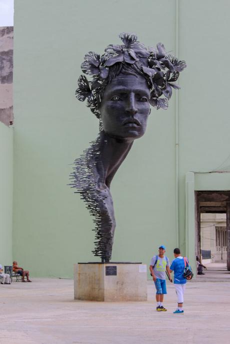 A modern sculpture of a fragmented head with floral adornments, displayed against a pale green wall, with two people standing nearby.