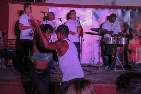 A lively scene of a couple dancing energetically on stage with a live band playing behind them. The musicians, wearing white shirts, play various instruments like drums and maracas, while the audience enjoys the vibrant performance.