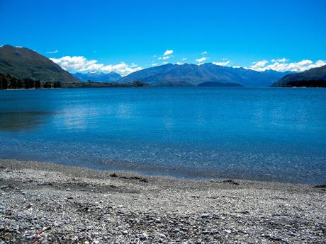 A clear blue lake with a pebble shoreline, surrounded by distant mountains under a bright blue sky.