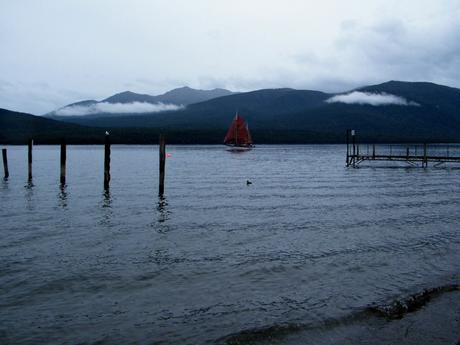 A lake with a small dock and wooden posts extending into the water, with a red-sailed boat in the distance against a backdrop of misty mountains.