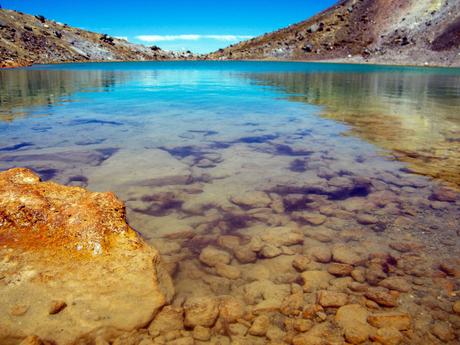 A close-up of a clear turquoise lake with visible stones beneath the water, set between rocky shores under a bright blue sky.