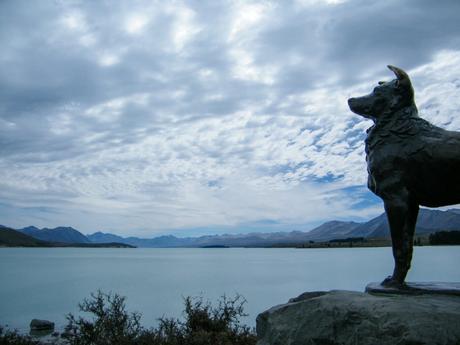 A bronze statue of a dog stands on a rock overlooking a lake with mountainous terrain in the background under a cloudy sky.