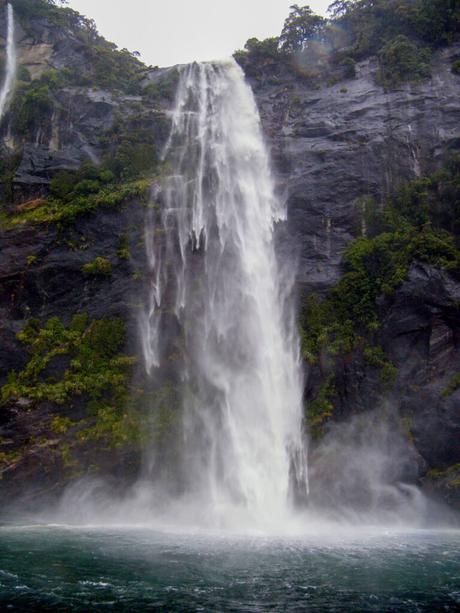 A powerful waterfall cascading down a steep cliff, surrounded by lush green vegetation, with mist rising from the pool of water at the base.