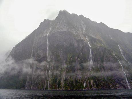 A towering, mist-covered mountain with multiple waterfalls cascading down its steep, dark cliffs into a fjord below.