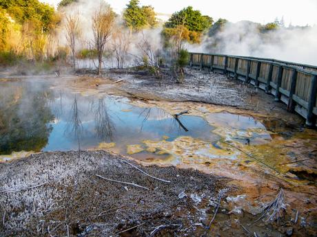 A geothermal hot spring surrounded by barren land, with mineral deposits and steam rising from the surface, and a wooden boardwalk in the background.