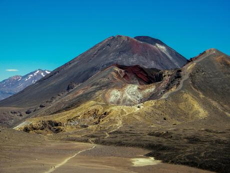 A rugged mountain landscape under a clear blue sky, featuring a volcanic peak with red and brown hues, surrounded by rocky terrain with sparse vegetation.