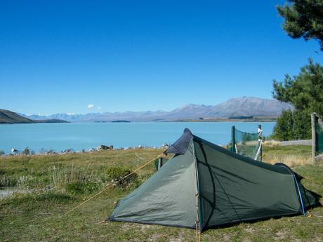 A camping tent pitched on grassy land near a pale blue lake, with distant mountains under a clear, sunny sky.