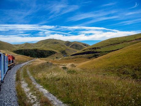 A train moving along a narrow, winding track through rolling grassy hills under a vibrant blue sky with wispy clouds.