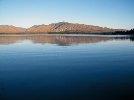 A calm lake reflecting the soft hues of a mountain range in the early morning light, under a pale blue sky.