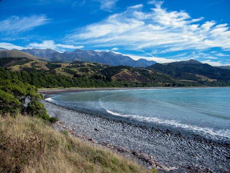 A scenic coastline with a pebble beach, bordered by green hills and mountains under a partly cloudy sky.